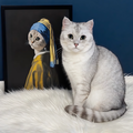 A cat sits beside a framed oil-style portrait of a feline wearing a blue headscarf, inspired by Girl with a