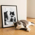 A framed print of a grey and white cat in a tie looking into a bathroom mirror, displayed in a